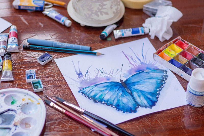 Photo d'un dessin d'un papillon bleu en cours de création, entouré de pinceaux et peinture acryliques (blue butterfly painted in watercolor). Copyright photo : Elena Mozhvilo https://unsplash.com/@miracleday